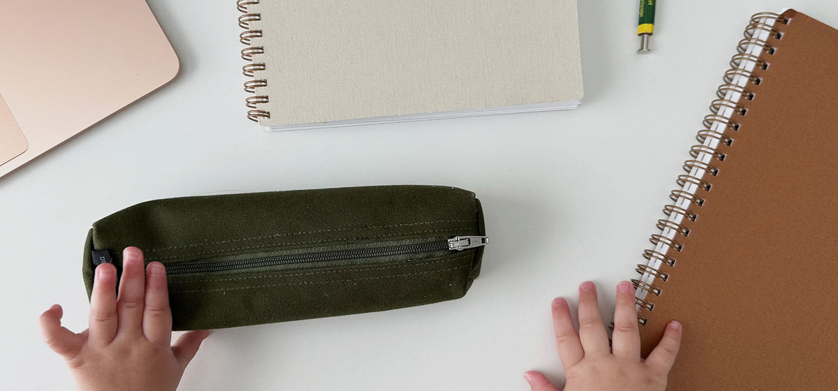 Appointed planners, workbooks, and a pencil case are strewn across a white table top. A baby's hand reaches out to grasp the pen case and planner.
