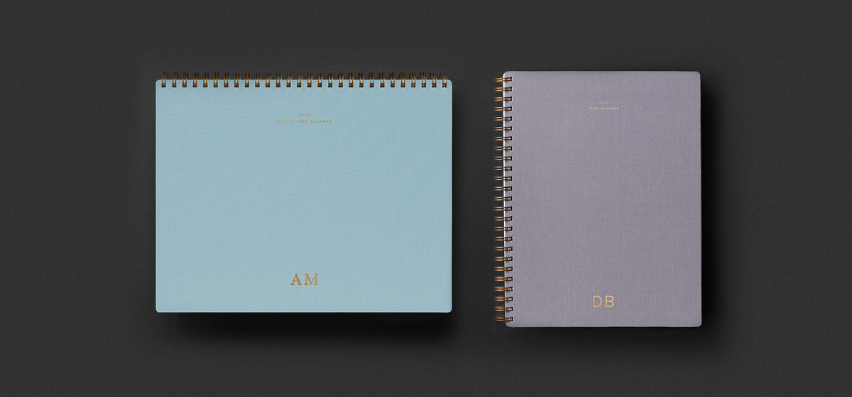 The Weekly Task Planner in Sky Blue and Compact Task Planner in Lavender Gray sit against a dark green background