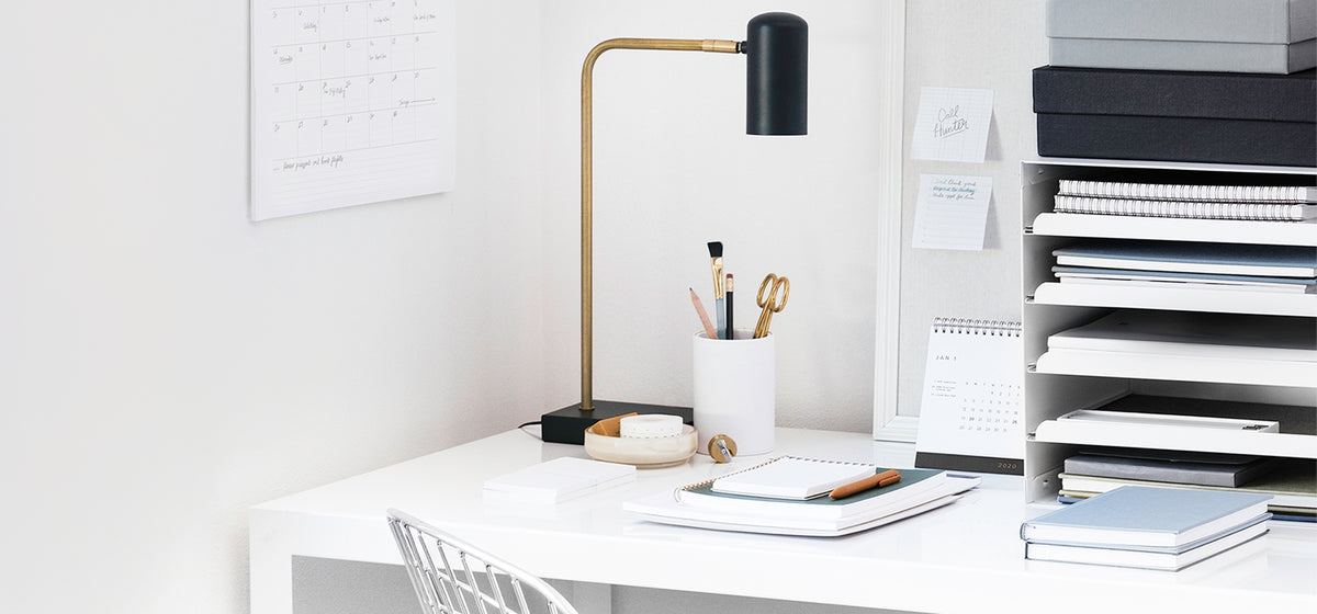 A serene, white workspace contains a white sek, black and gold desk lamp, vertical file organizer, various stationery items including desk accessories in a cup and a light blue hardcover books, as well as the bottom of a wall calendar.