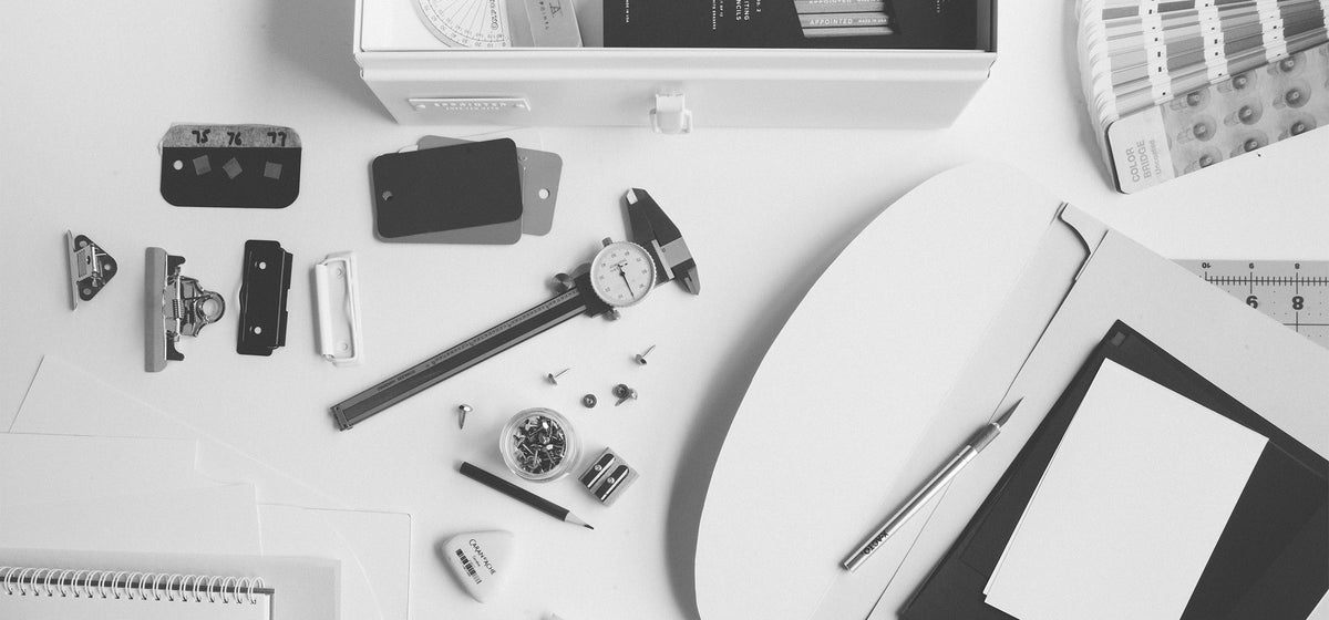 A white desk with various product design tools scattered about, including measuring tools, clips, push pins, swatch cards, pencils, and an exacto knife.The image is in black and white.